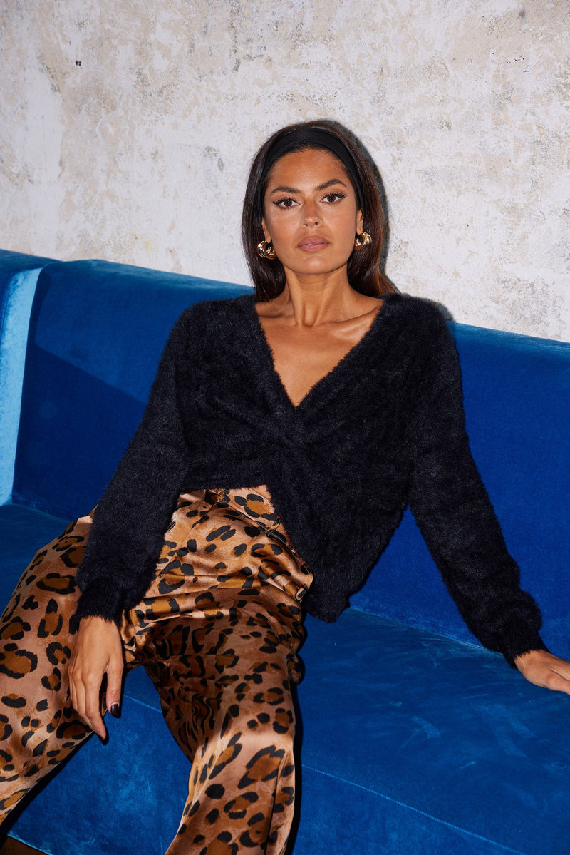 Female model sitting down. She wears a Dancing Leopard black jumper with a tie-knot front and leopard print trousers.