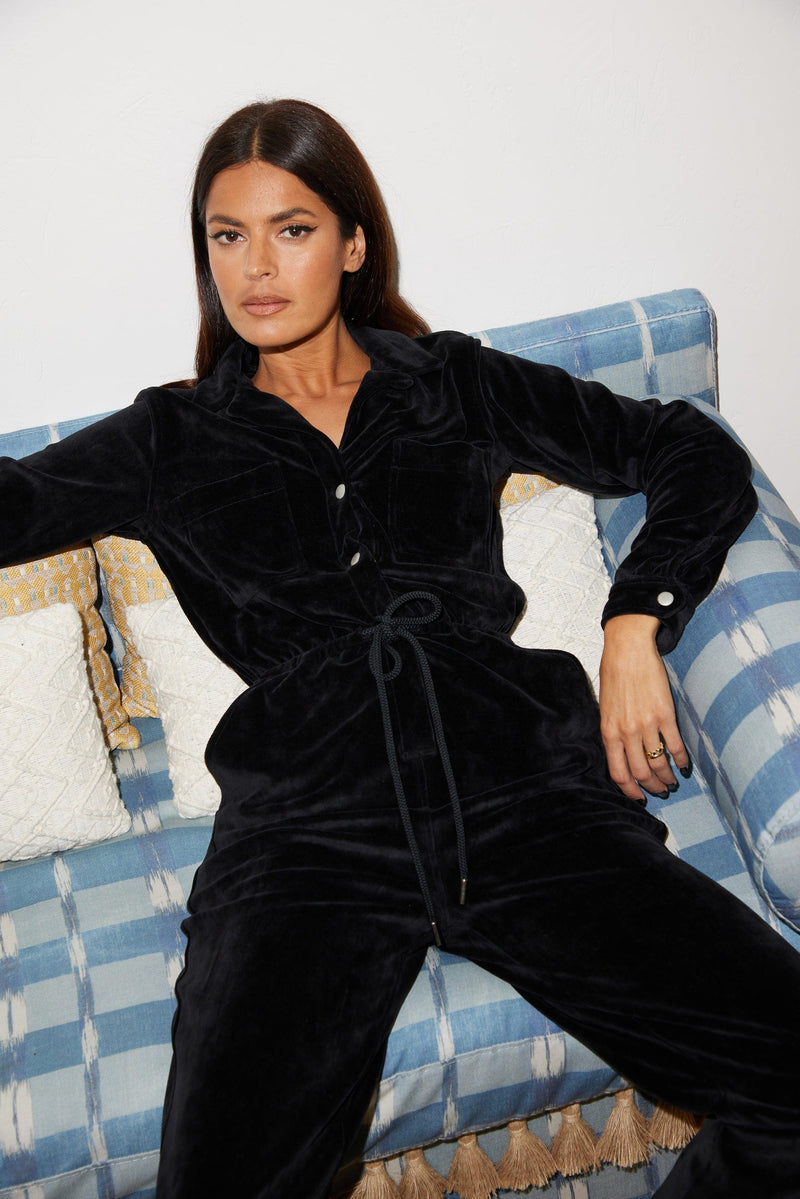 Female model sitting down on a couch. She wears a Dancing Leopard black velour jumpsuit.